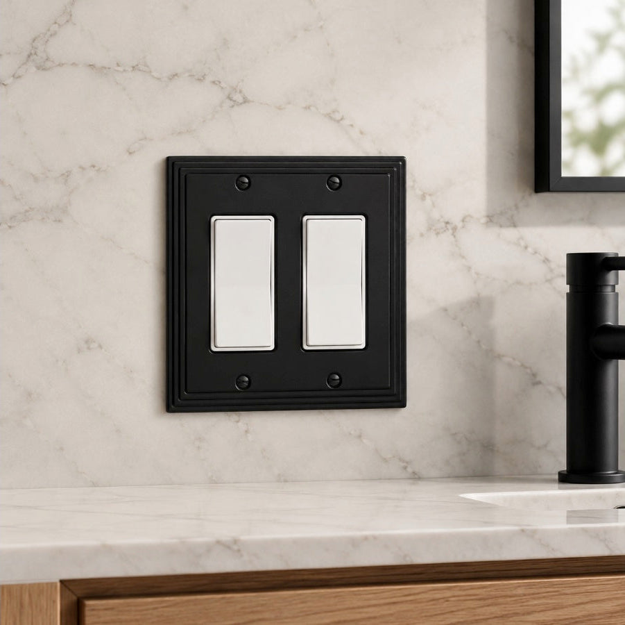 A black double light switch plate cover mounted on a white marble bathroom wall above a marble countertop with a black faucet.