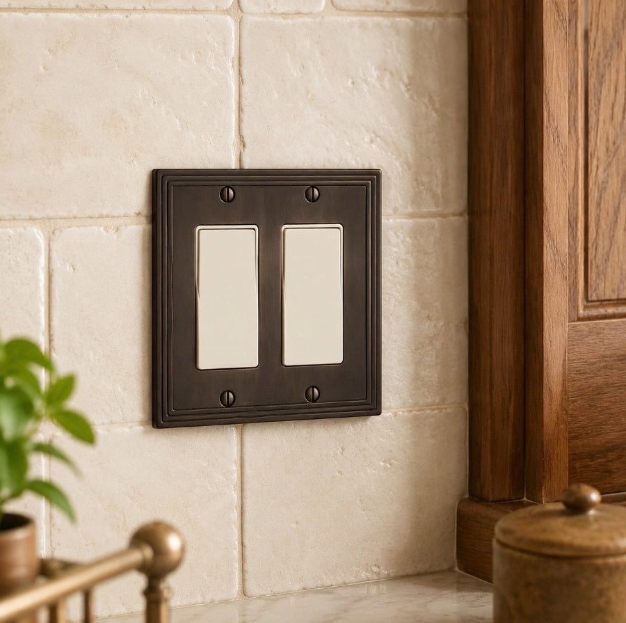 A double-gang light switch plate set in a dark oil-rubbed bronze finish, mounted on a beige stone tile wall next to a wooden cabinet.