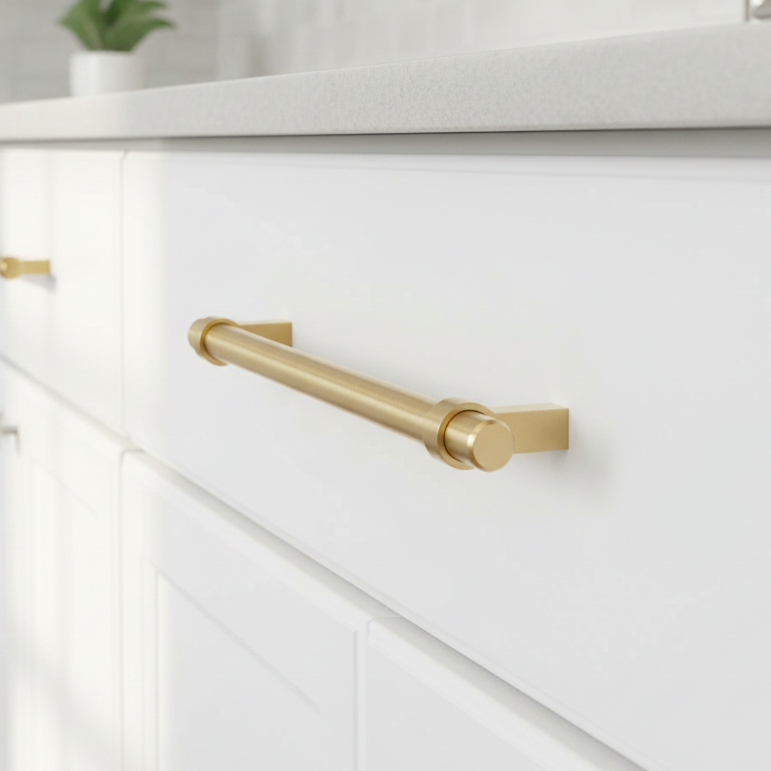 A Cosmas brushed gold drawer pull handle mounted on a white kitchen cabinet drawer, with a light countertop and a small blurred plant in the background.