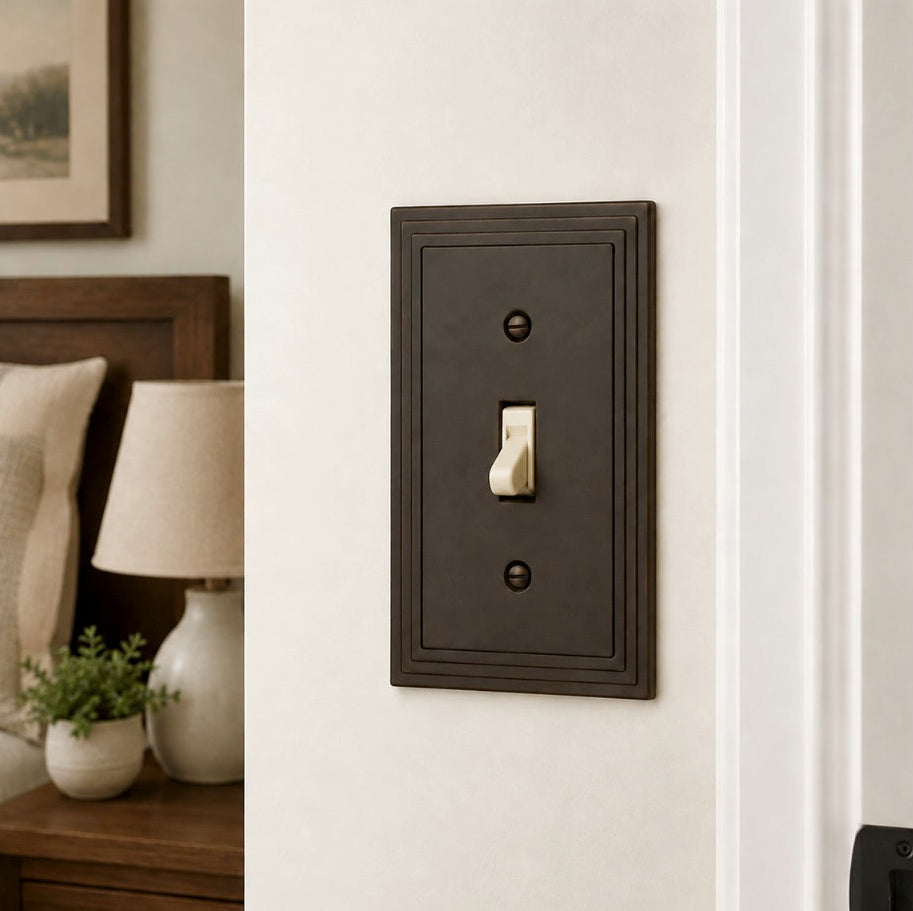 A single-toggle light switch plate in a dark bronze finish with a cream-colored lever switch, mounted on a white wall in a bedroom setting.