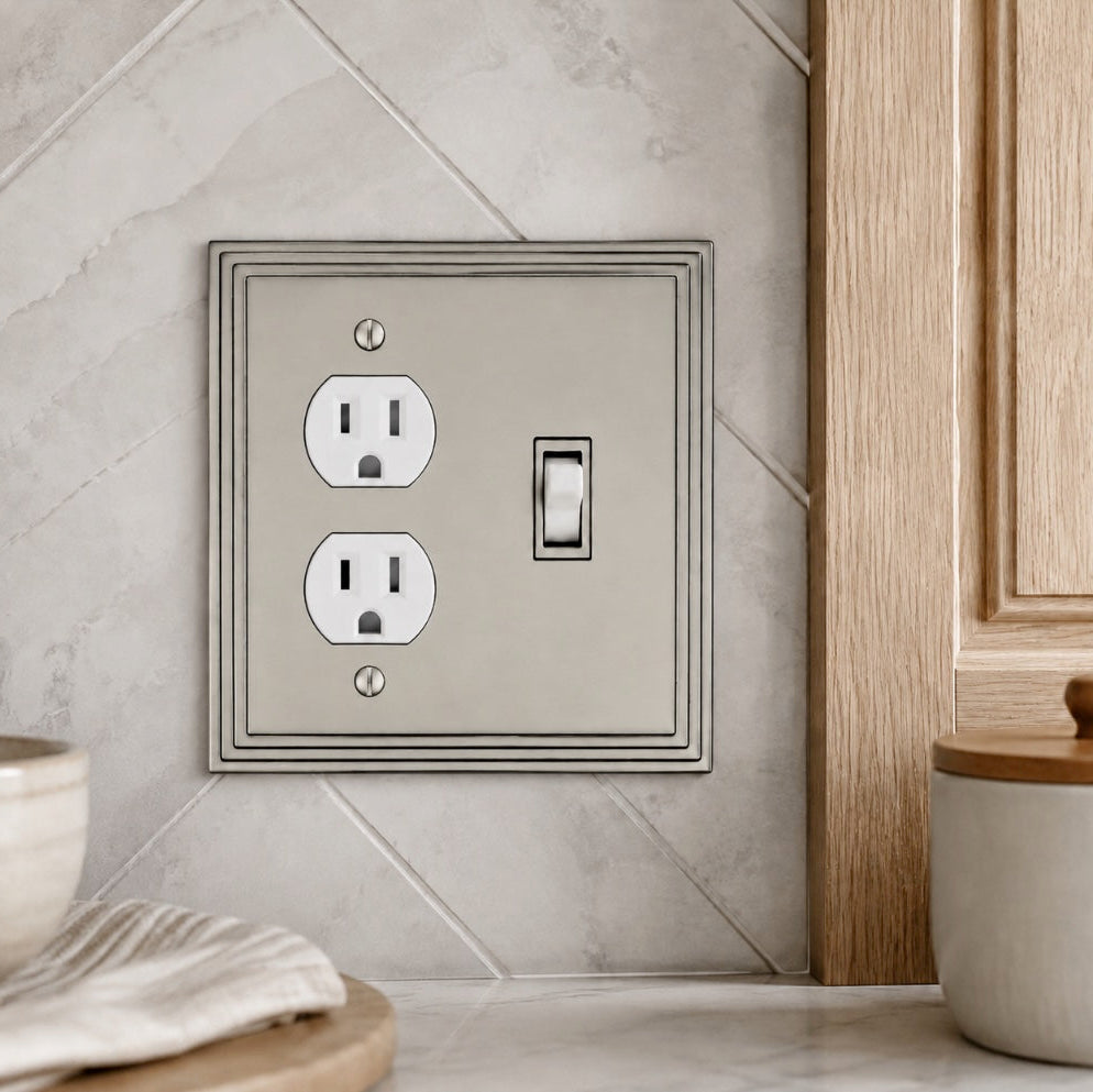 A decorative wall plate featuring a single toggle switch and two electrical outlets, styled in a kitchen setting with a tiled backsplash and wooden cabinetry.